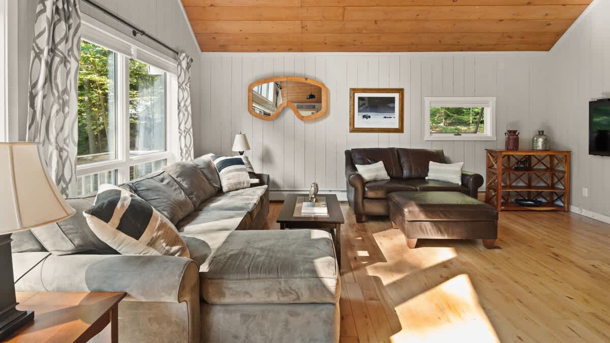 Spacious living room with light wood floors and large windows, furnished with a gray sectional sofa and a brown leather armchair with an ottoman. A unique ski goggle-shaped mirror adorns the light gray shiplap wall.