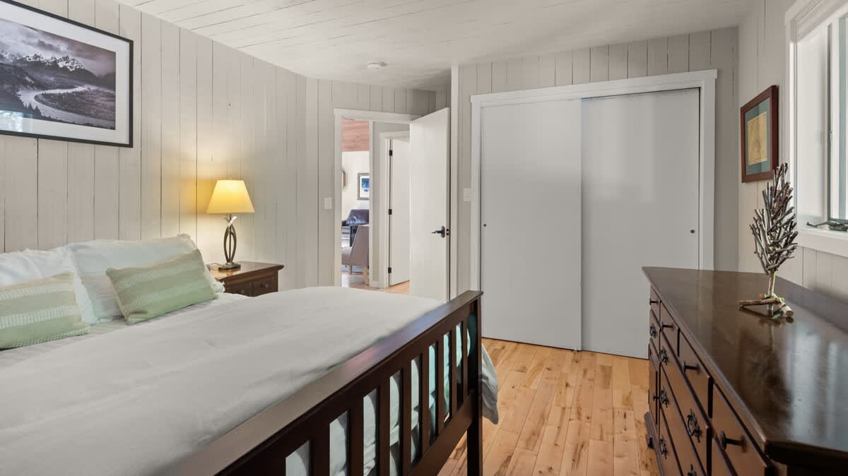 A bright bedroom with light wood floors and white shiplap walls, featuring a dark wooden bed frame and a nightstand with a lamp. Sliding closet doors and a doorway leading to another room are visible.