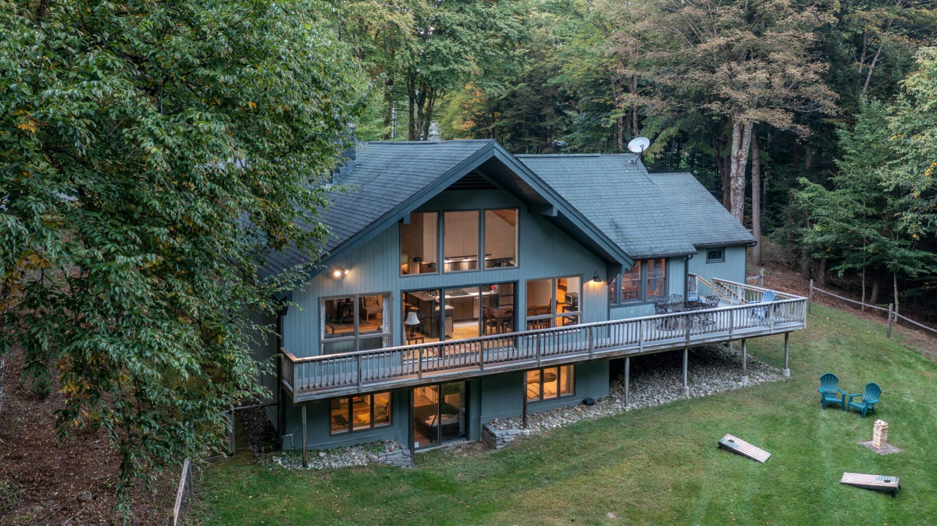 Aerial view of a large, modern chalet-style house with a dark blue-green exterior, multiple large windows, and a long wooden deck, nestled among lush green trees on a grassy slope.