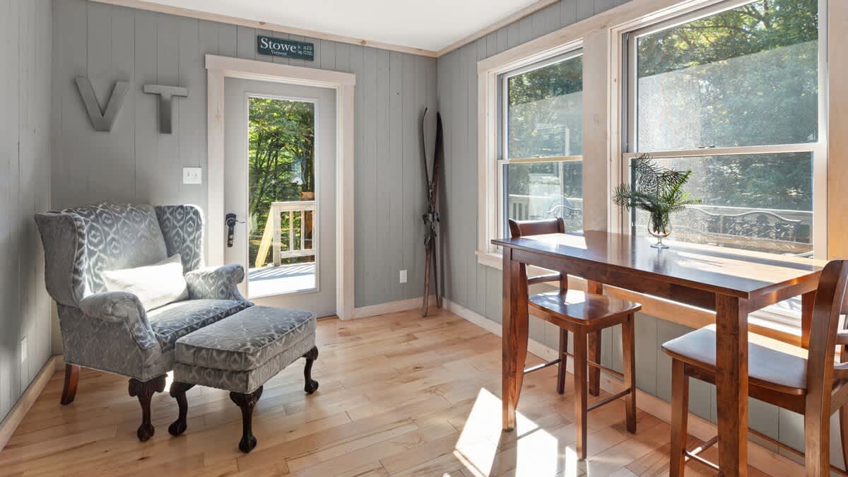 Inviting sunroom or secondary sitting area featuring a comfortable gray upholstered armchair and ottoman, alongside a high-top wooden table with bar stools. The room is well-lit by large windows and has light wood paneling and hardwood floors.
