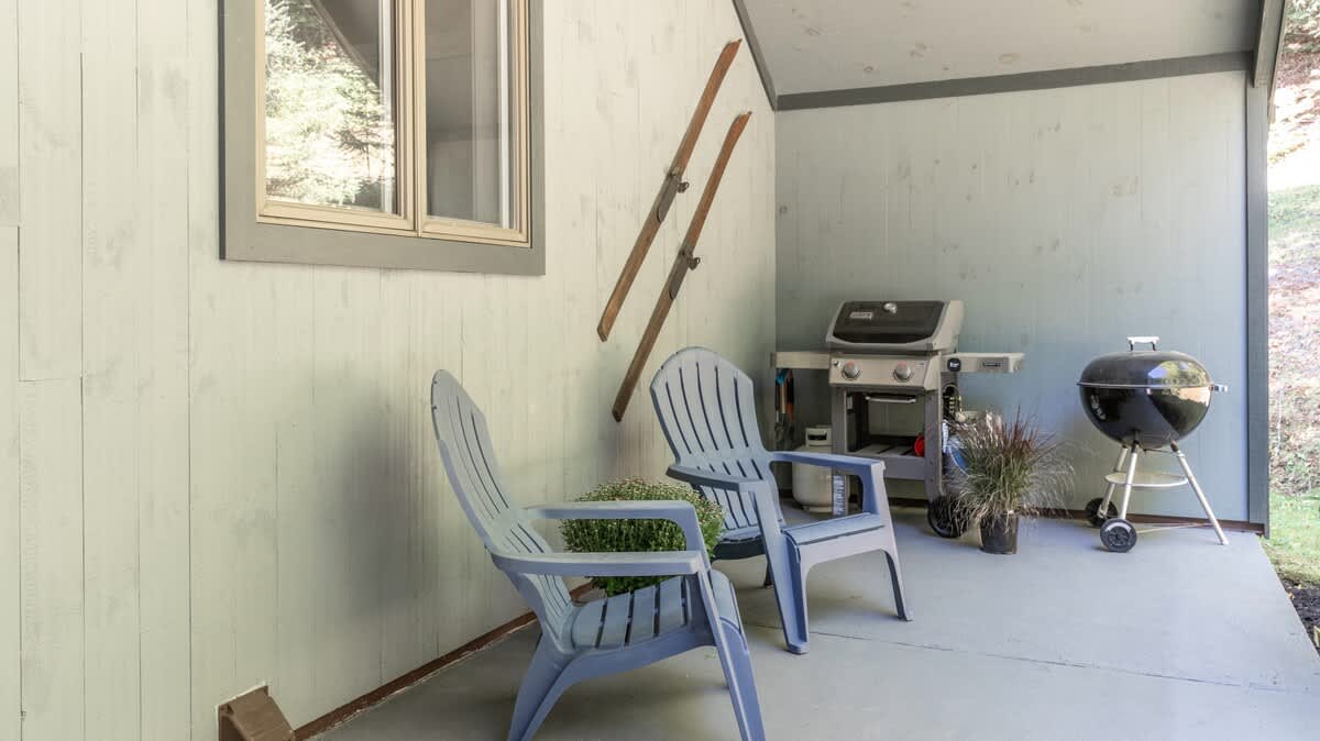 Covered outdoor patio area featuring two light blue Adirondack chairs, a gas grill, and a charcoal grill, with vintage skis mounted on the light blue shiplap wall.
