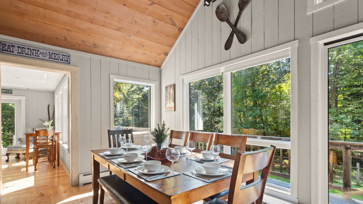 Inviting dining room with a wooden table, bench, and chairs, set against a backdrop of large windows showcasing the surrounding forest. The room features light wood paneling, a vaulted wood ceiling, and a doorway leading to another bright space.