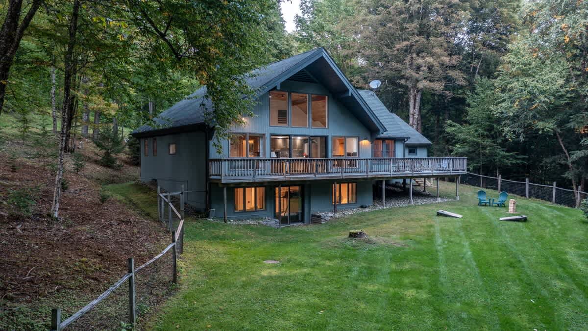 A modern chalet-style house with a dark blue-green exterior and a large wooden deck, nestled on a grassy slope surrounded by dense green trees. The house features large windows and a walk-out lower level.