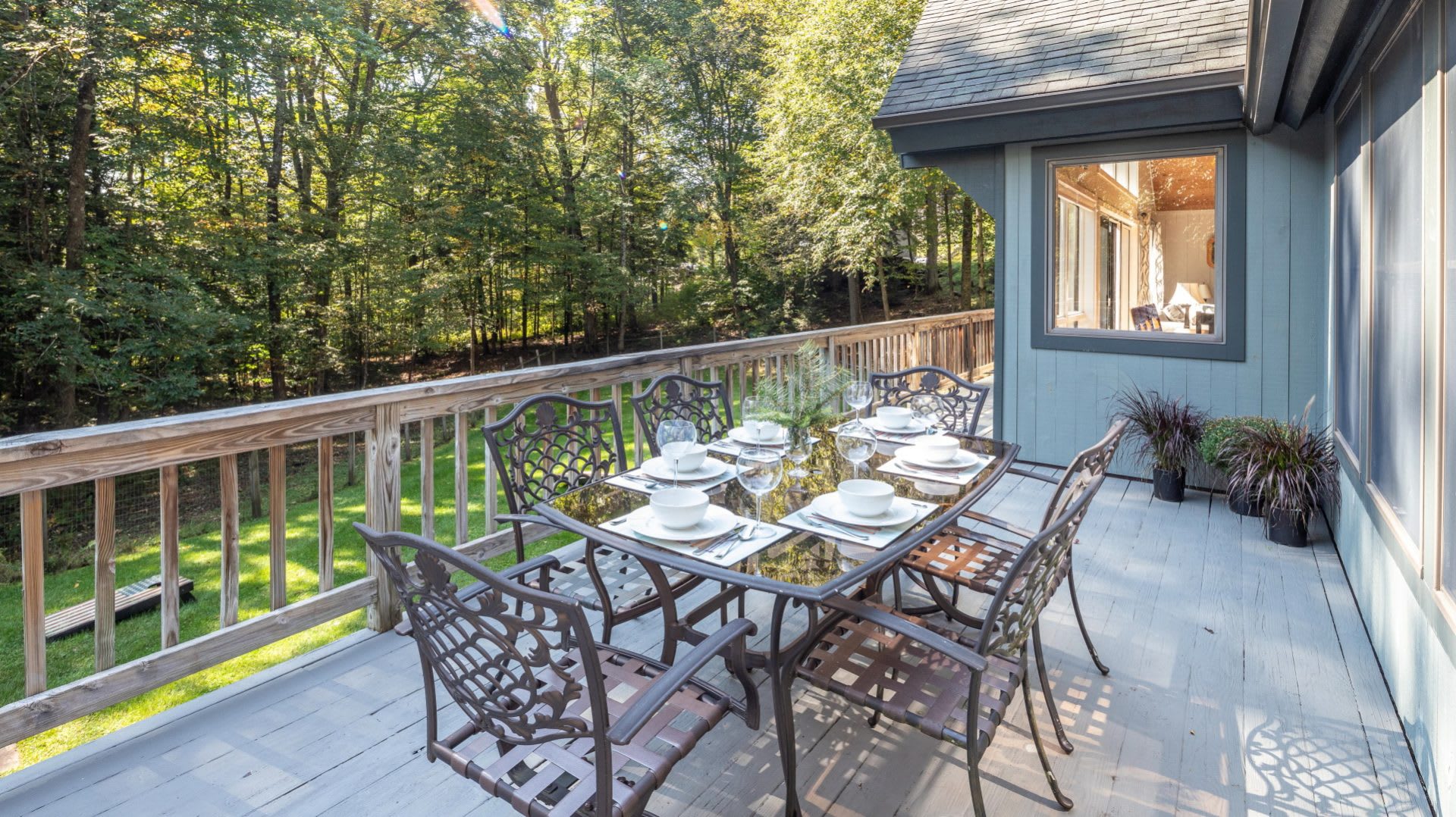 Spacious outdoor deck with a wrought iron dining set for six, complete with place settings, offering a scenic view of the lush green backyard and dense surrounding forest.
