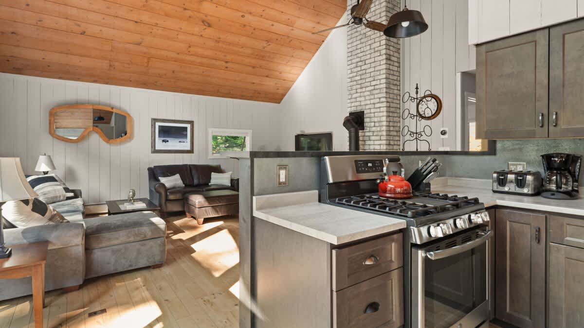 Modern kitchen area with stainless steel appliances, including a gas range and oven, and dark wood cabinetry. A breakfast bar separates the kitchen from the open-concept living area, which features a vaulted wood ceiling and light wood floors.