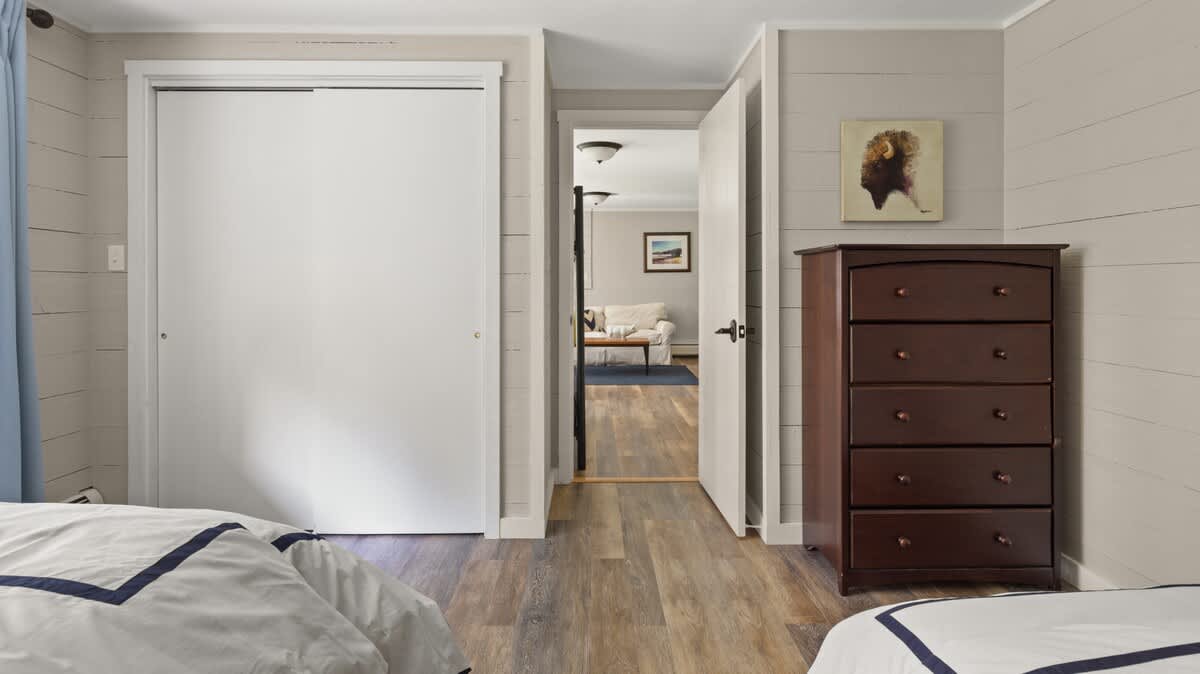 A bedroom with two twin beds, dark wood-look flooring, and light gray shiplap walls, featuring a tall wooden dresser. Sliding closet doors and a doorway leading to another room are visible, enhancing the functional layout.