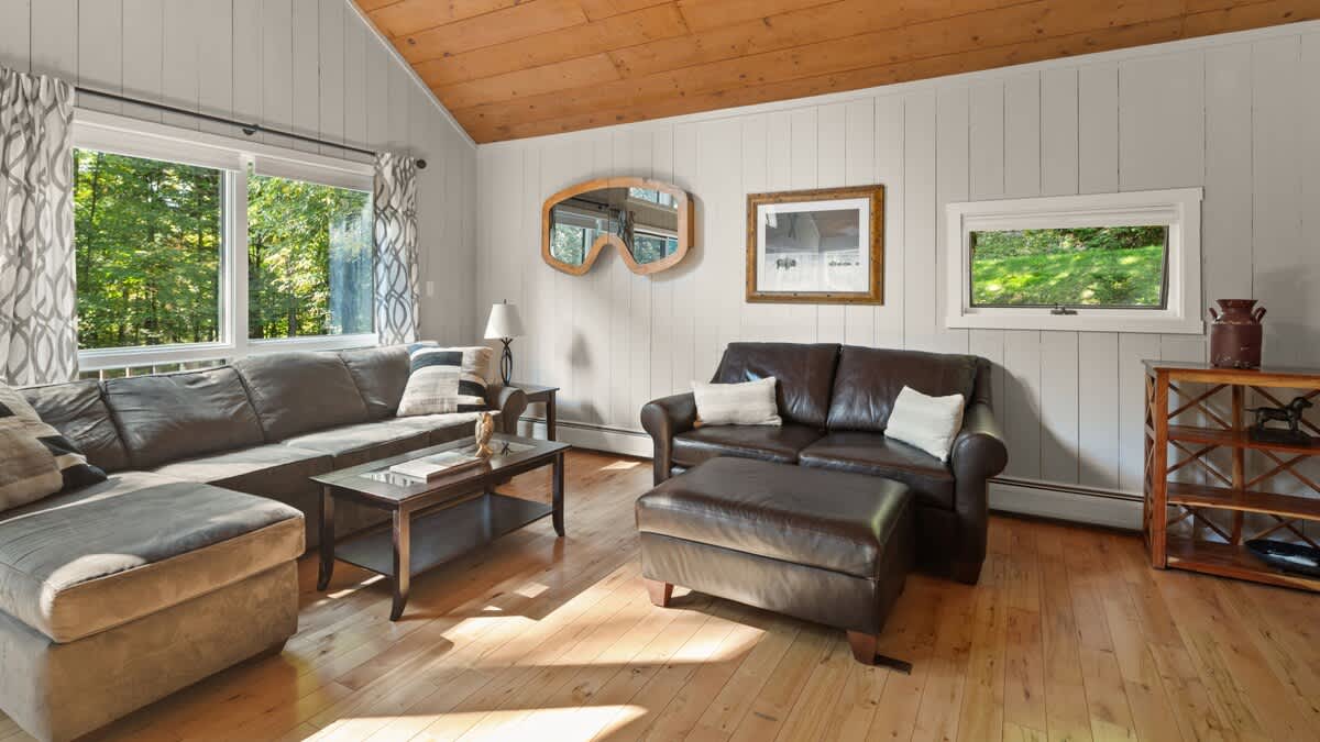 Spacious living room with a brown sectional sofa, a matching leather armchair and ottoman, and a glass-top coffee table. Light wood floors, light wood paneling, and a large window offering a view of the surrounding greenery complete the cozy space.