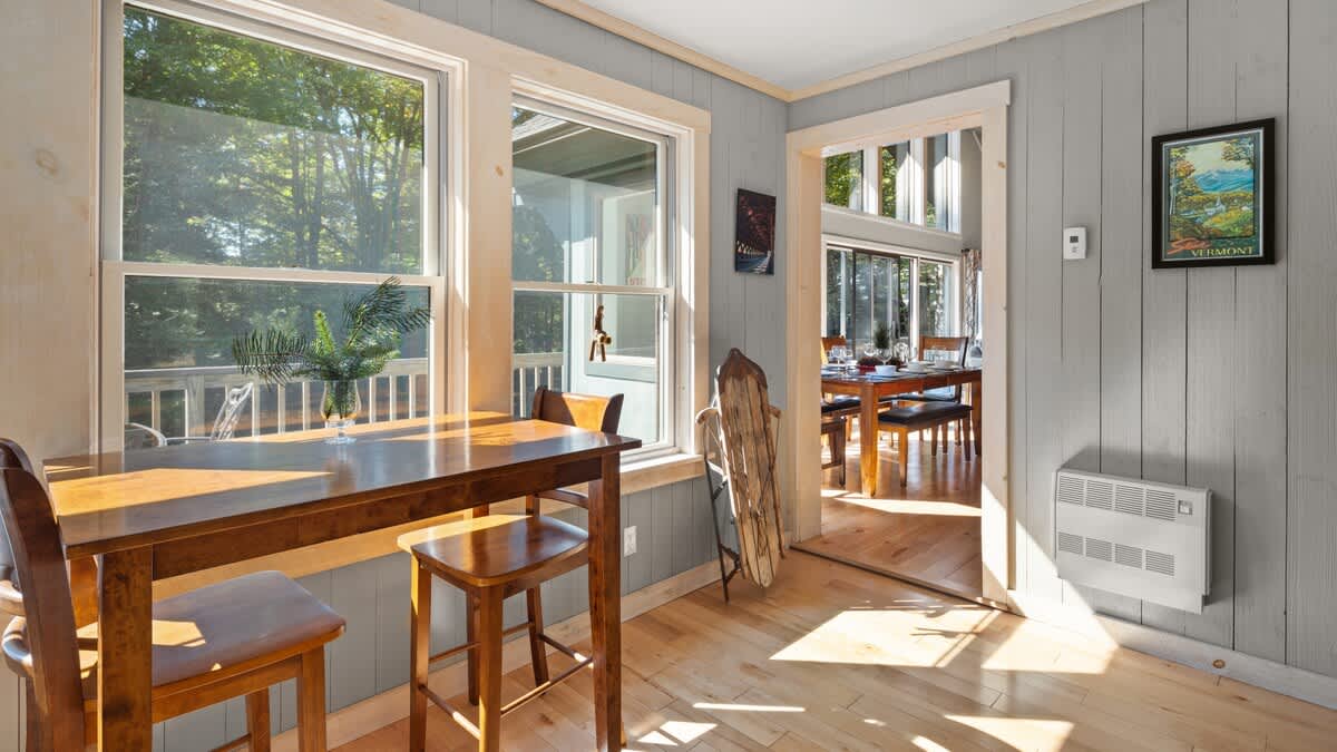 Bright sunroom or breakfast nook with a high-top wooden table and two bar stools, surrounded by large windows offering views of the outdoors. The room features light wood paneling and hardwood floors, with a doorway leading to the main dining area.