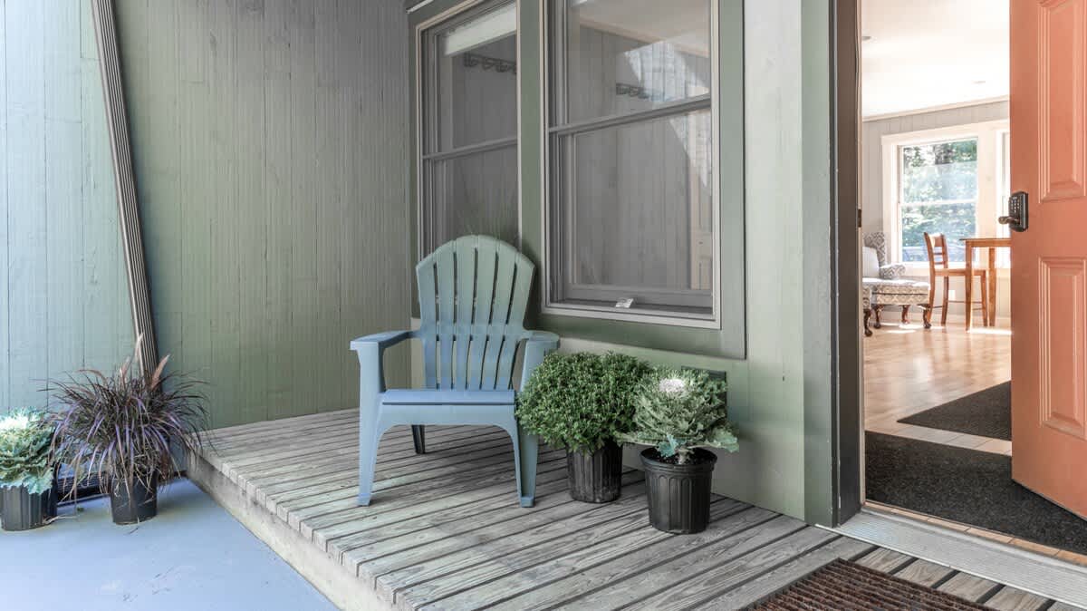 Inviting front porch with a light blue Adirondack chair, potted greenery, and a warm orange front door with a keyless entry pad, set against a light green exterior wall.