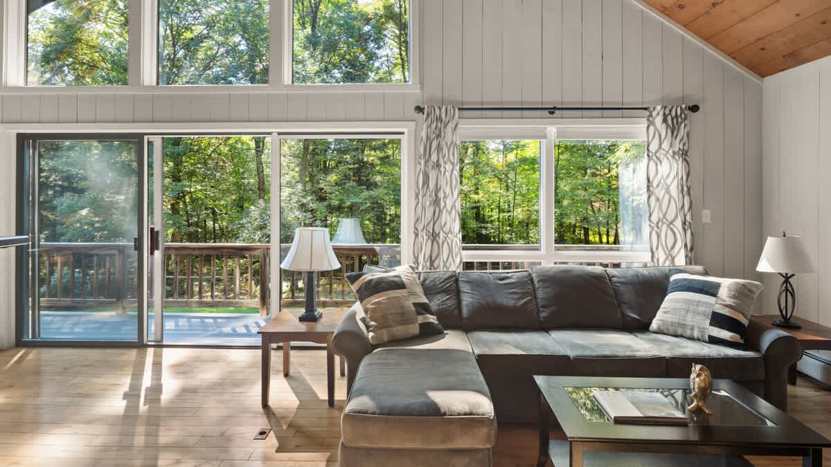 Bright living room featuring a large brown sectional sofa with decorative pillows, a glass-top coffee table, and expansive windows and a sliding glass door leading to a deck with forest views. The room has light wood floors and a vaulted wood ceiling.
