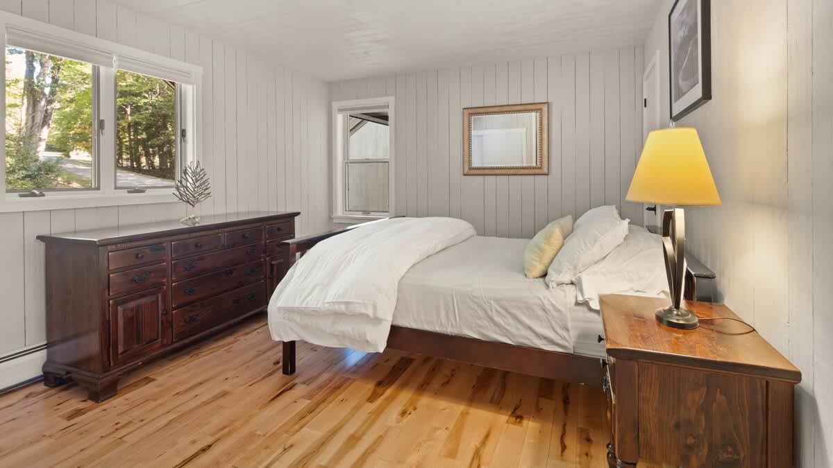 A serene bedroom with light wood floors and white shiplap walls, featuring a dark wooden bed frame and a matching dresser. A nightstand with a lamp and a window overlooking greenery complete the inviting space.