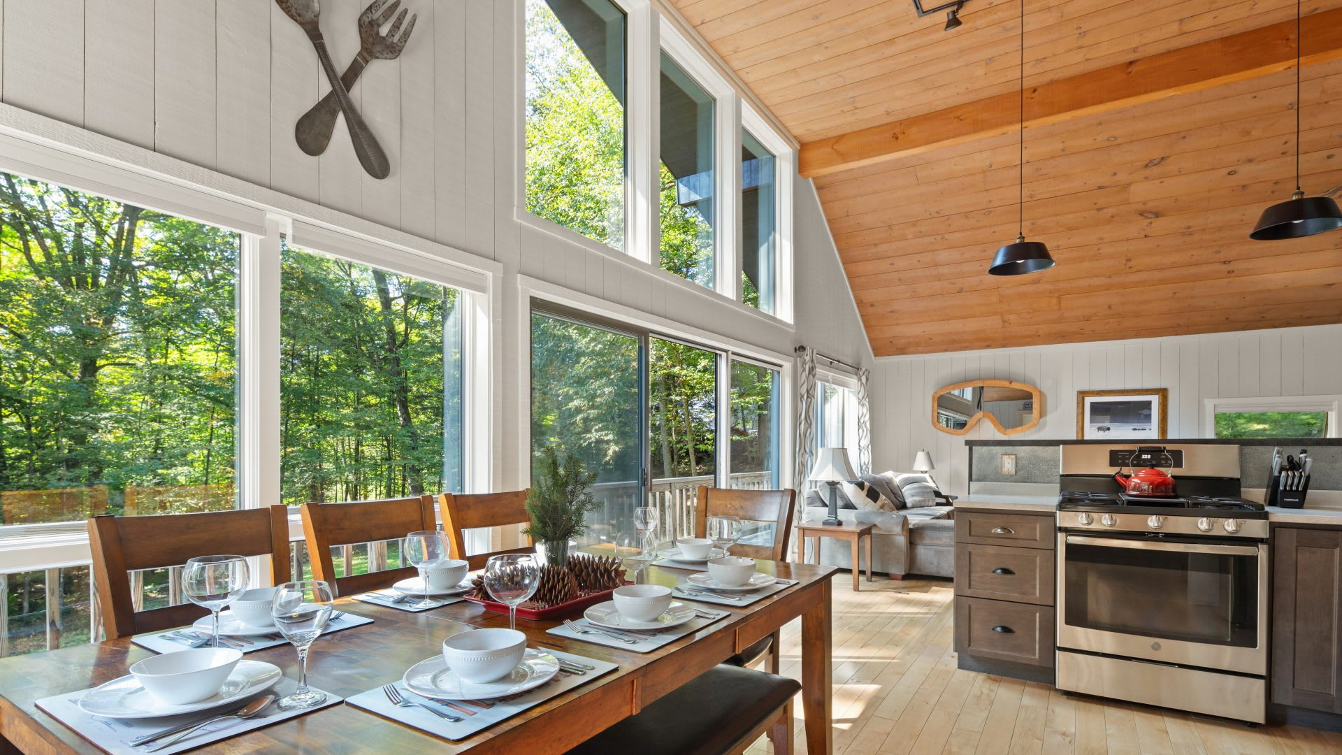 Elegant dining area with a long wooden table set for six, surrounded by large windows that provide abundant natural light and views of the lush forest. The space connects to the living room and kitchen, featuring a vaulted wood ceiling and decorative wall art.