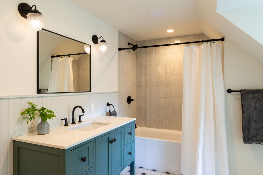 A modern bathroom featuring a teal vanity with a white countertop and black hardware, a rectangular mirror, and a shower/tub combo with white subway tile and black fixtures. The floor has a black and white patterned tile.