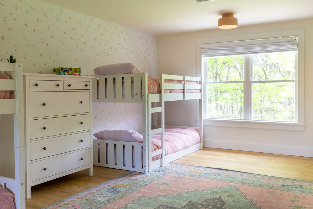 A bright children's bedroom featuring white bunk beds with pink bedding, a matching white dresser, and a light-colored rug with pink and blue patterns. The room has light wood floors and a large window overlooking lush greenery.