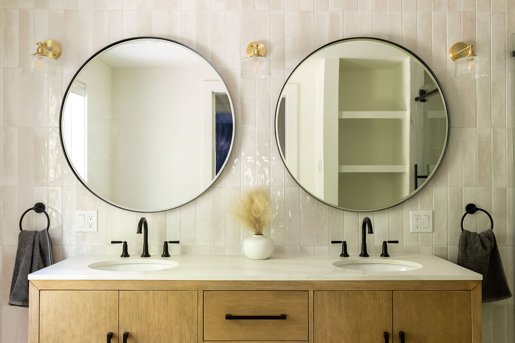 Double vanity bathroom featuring two round black-framed mirrors, black faucets, and a light wood cabinet with ample storage. The wall is adorned with vertical textured white tiles, and brass sconces provide lighting.