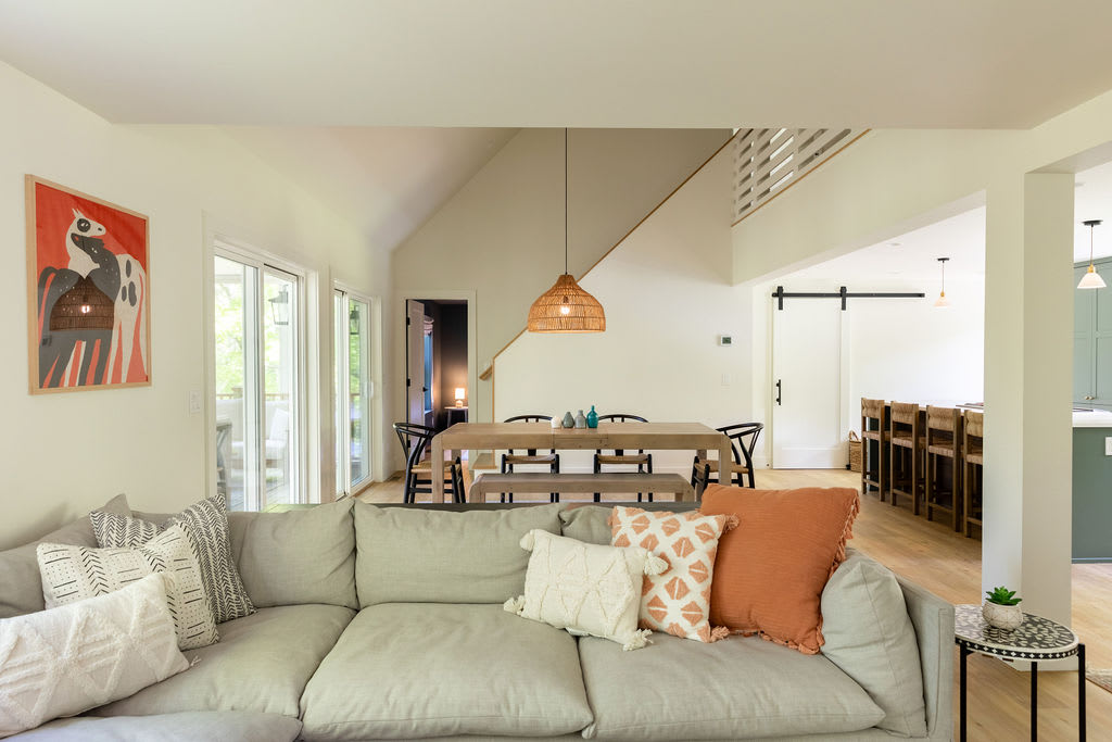 An open-concept view from the living room towards the dining area and kitchen, featuring a light gray sectional sofa and a glimpse of the dining table. The space has light wood floors, white walls, and a high ceiling with a staircase visible in the background.