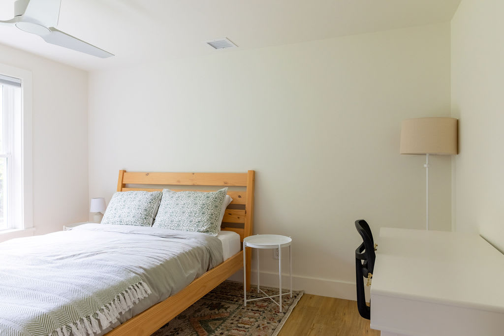 A wider view of the serene bedroom, featuring a light wood bed with grey bedding, a small white side table, and a white floor lamp. The room has light wood floors and a large window, with a simple rug under the bed.