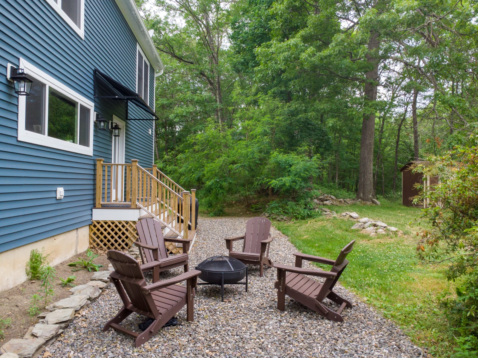 An outdoor seating area with four dark brown Adirondack chairs arranged around a black metal fire pit, set on a gravel patio next to the blue house. Lush green trees and foliage provide a natural backdrop.