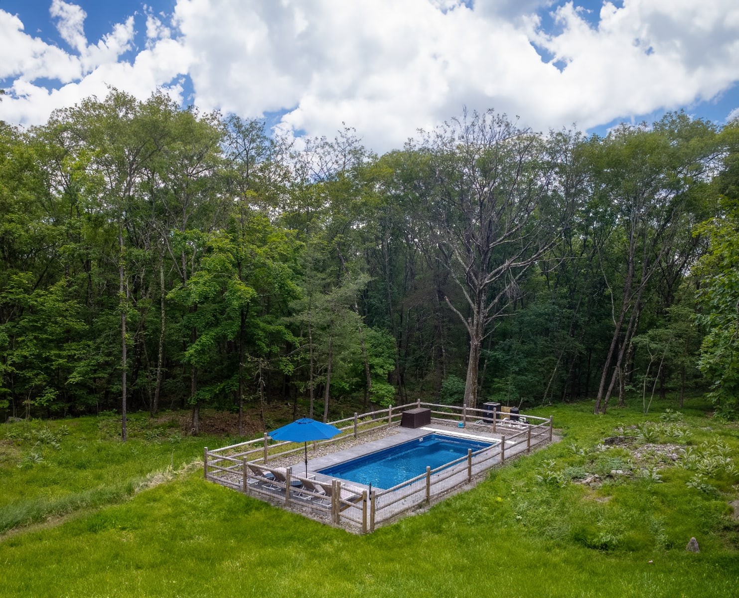 An aerial view showcases a rectangular outdoor swimming pool with a blue umbrella and lounge chairs, enclosed by a wooden fence, nestled in a grassy clearing surrounded by dense green forest.