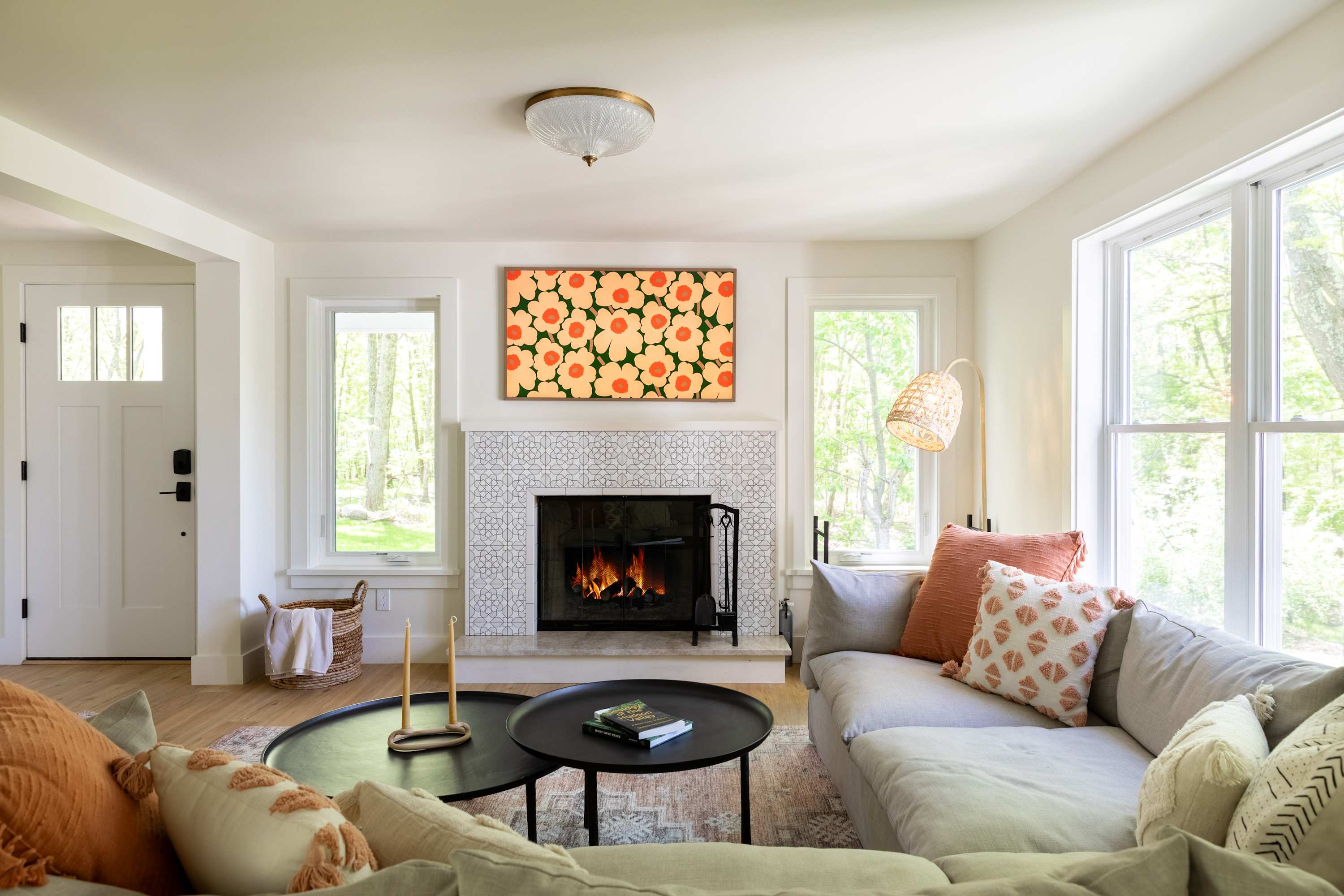 A bright living room with a large L-shaped sectional sofa adorned with orange and white throw pillows, facing a tiled fireplace with a modern art piece above it. Large windows offer views of the surrounding greenery, and two black coffee tables sit on an area rug.