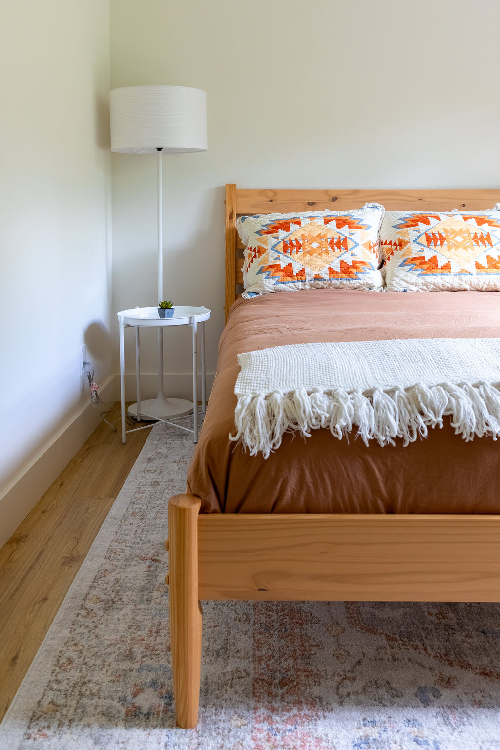A close-up view of a light wood bed frame with brown bedding and two decorative pillows featuring orange, blue, and white geometric patterns. A white floor lamp and a small white round side table with a plant are visible beside the bed.