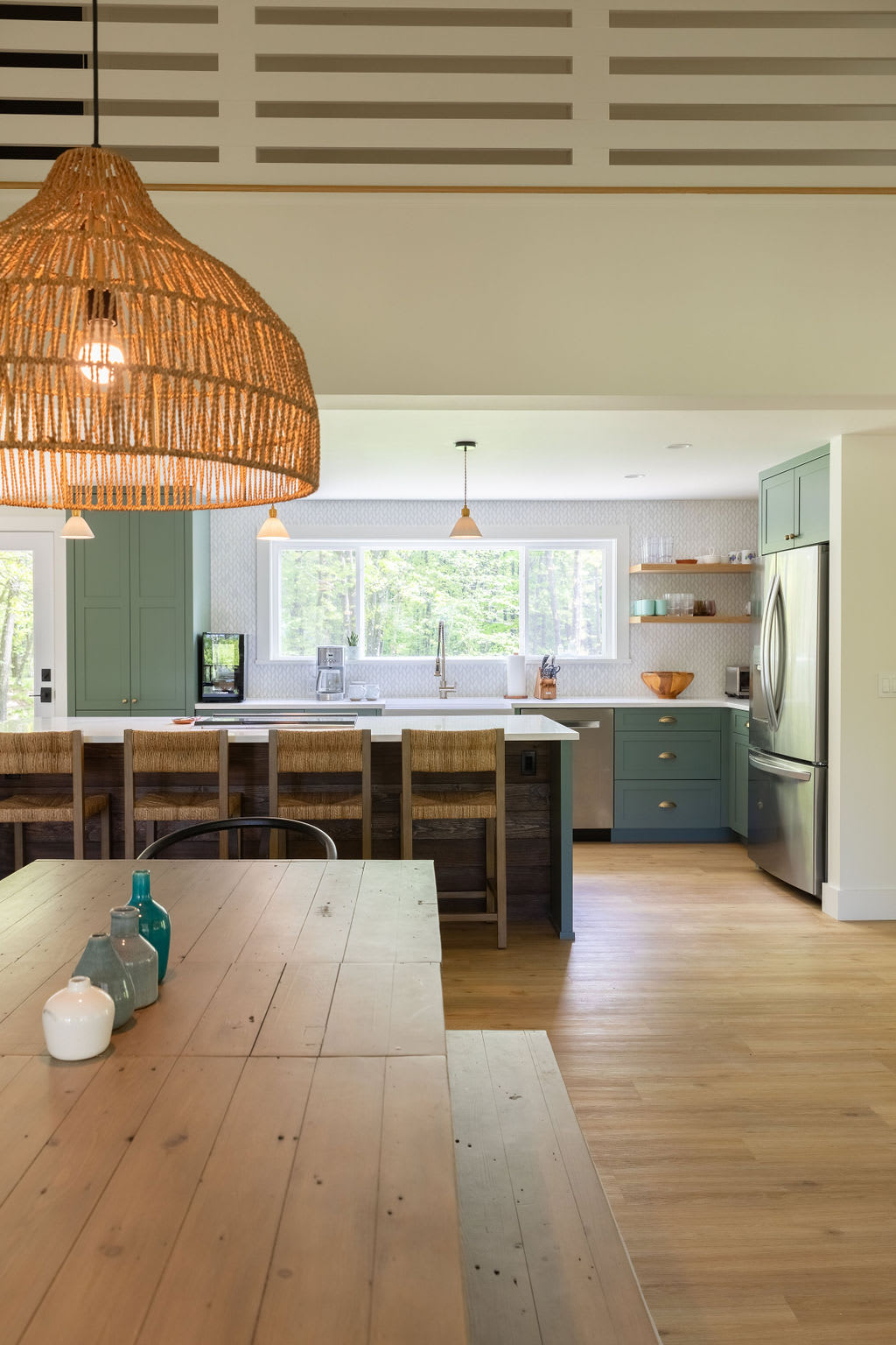 Open-concept kitchen and dining area featuring sage green cabinetry, a large island with bar seating, and a rustic wooden dining table under a woven pendant light. Large windows overlook a lush green forest.