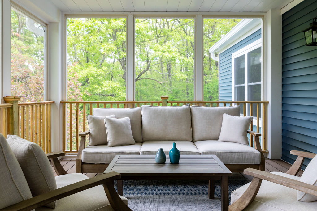 A comfortable screened porch furnished with a light-colored outdoor sofa and armchairs, centered around a dark wood coffee table. The porch overlooks a lush green wooded area through large screened windows.