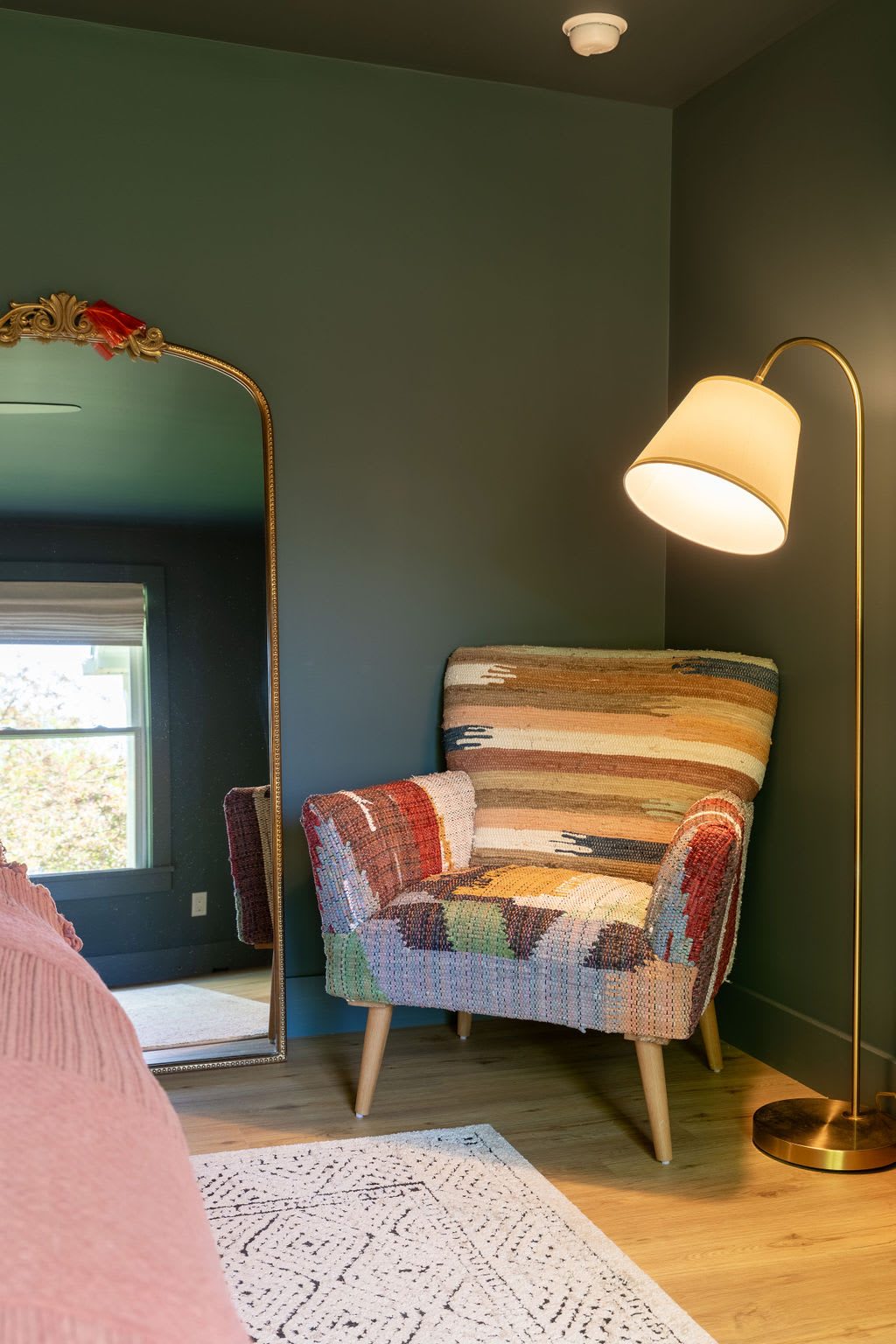 Corner of a bedroom with a vibrant, patterned armchair and a tall brass floor lamp, complementing the dark green walls. A large gold-framed mirror leans against the wall, reflecting a window.