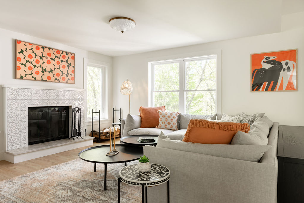 A bright living room features a large light gray sectional sofa with colorful throw pillows, centered around two black round coffee tables on a patterned rug. A white tiled fireplace with a black screen and a large abstract artwork adorns one wall.