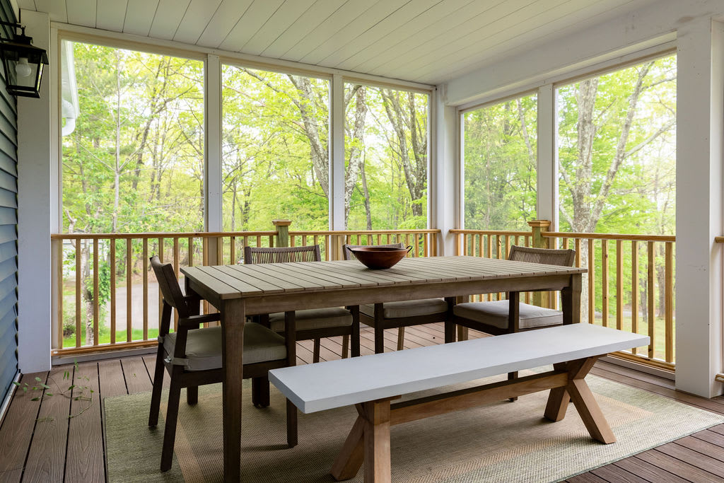 Another view of the screened-in porch, featuring a wooden dining table with chairs and a bench, set on a patterned rug. The space is enclosed by large windows, offering bright views of the lush green foliage outside.