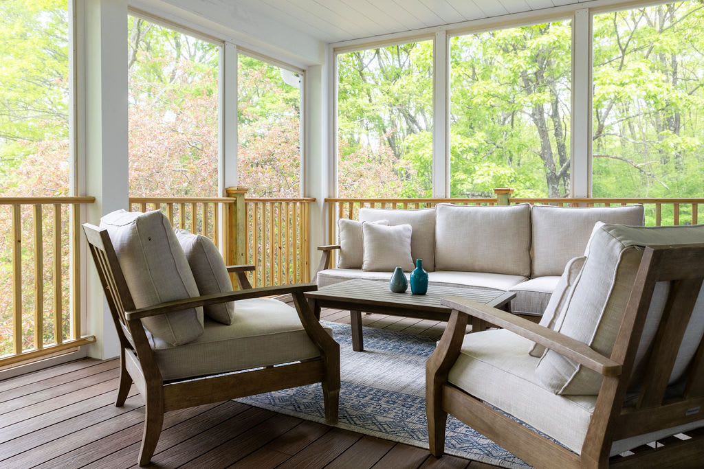 A screened-in porch offers a comfortable seating area with light-colored upholstered wooden chairs and a sofa, arranged around a low wooden coffee table. Large windows provide expansive views of the surrounding vibrant green trees.