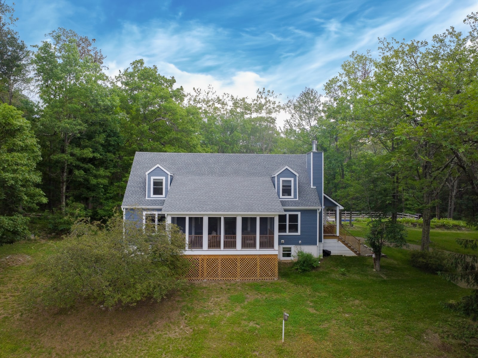 Aerial view of a blue two-story house with a gray roof and dormer windows, featuring a screened porch on the lower level, surrounded by a lush green lawn and dense forest.
