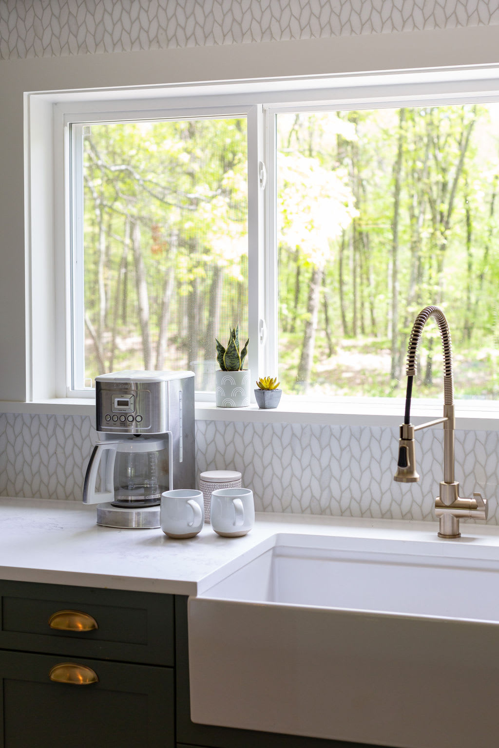 Detailed view of the kitchen sink area, showcasing a white farmhouse sink, a modern pull-down faucet, and a coffee maker on the white quartz countertop. A large window provides a bright view of the verdant trees outside.