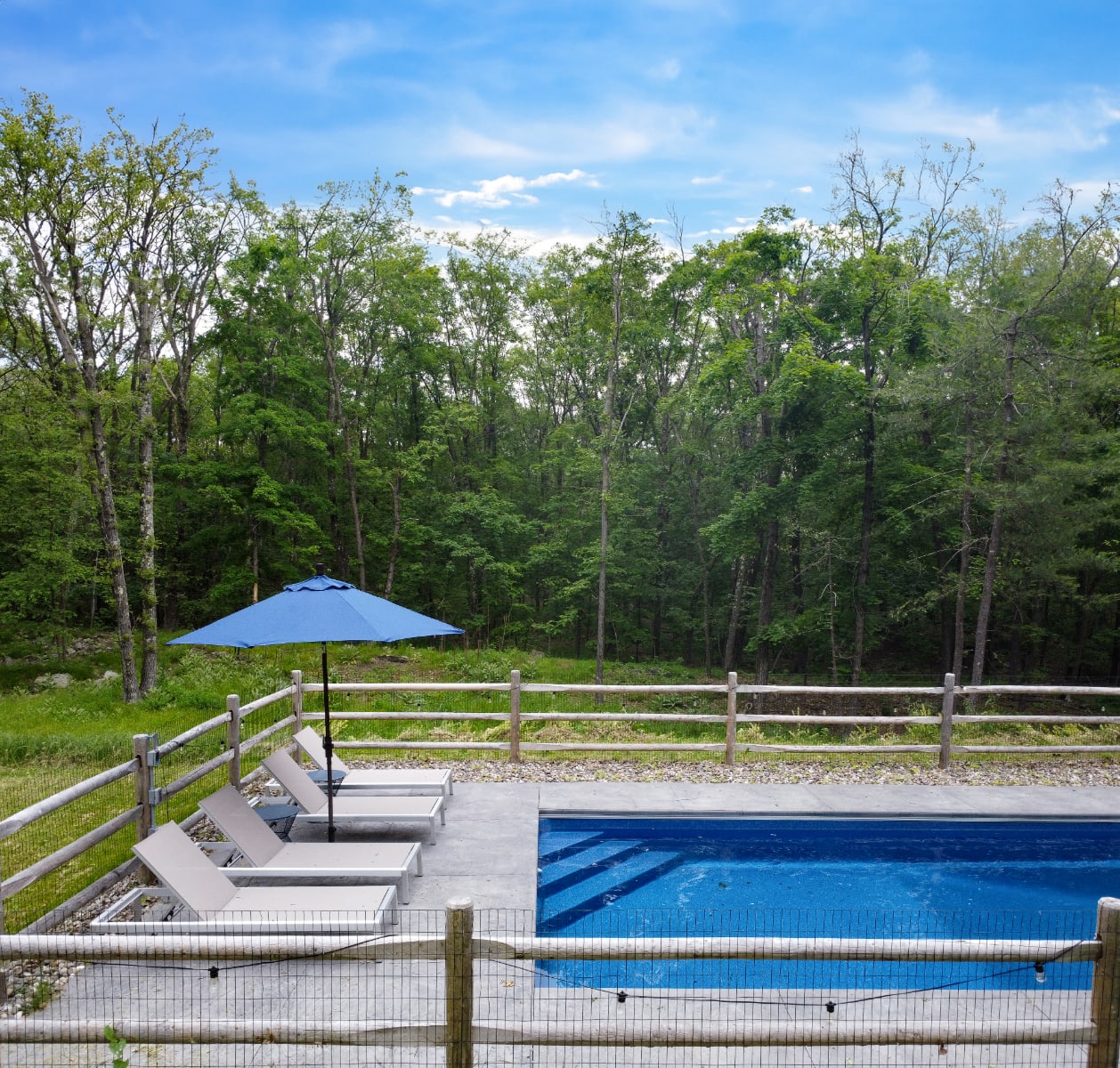 A ground-level view of a rectangular inground swimming pool with a concrete deck, three lounge chairs, and a blue umbrella, enclosed by a rustic wooden fence. The background features a dense forest under a bright blue sky.