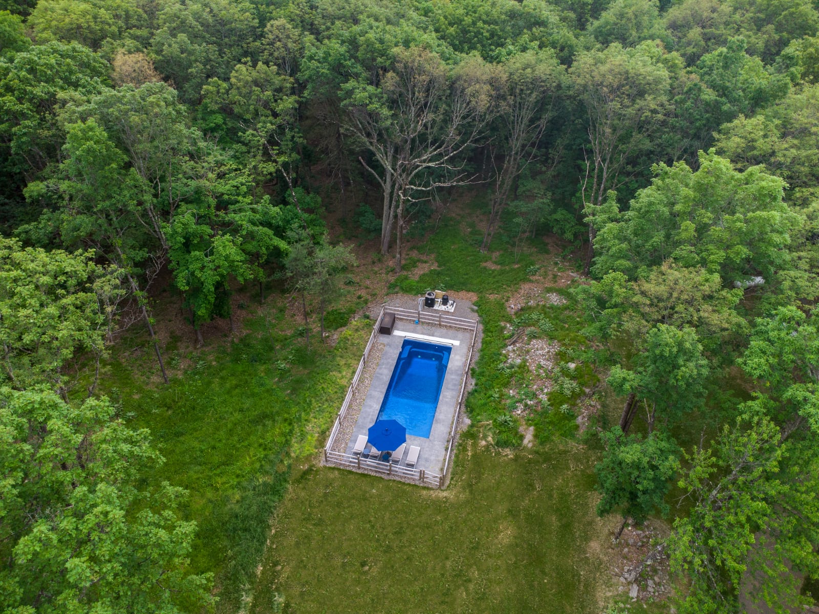 High aerial shot of a rectangular outdoor swimming pool with a blue umbrella, surrounded by a wooden fence and a grassy area, set within a vast, verdant forest under a cloudy sky.