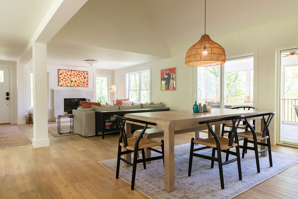 The dining area features a light wood rectangular dining table with black chairs and a bench, set on a light patterned rug. A woven pendant light hangs above the table, and large sliding glass doors lead to an outdoor porch.