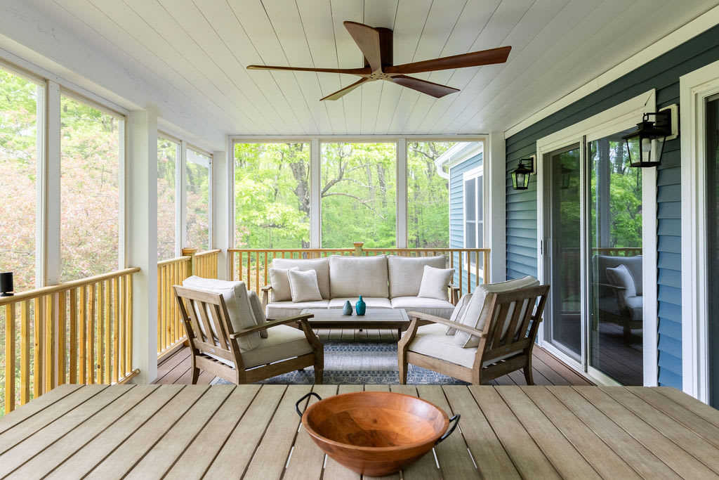 A spacious screened-in porch with a comfortable outdoor sofa and two armchairs, a coffee table, and a ceiling fan. The porch offers expansive views of the surrounding lush green trees, with a wooden dining table in the foreground.