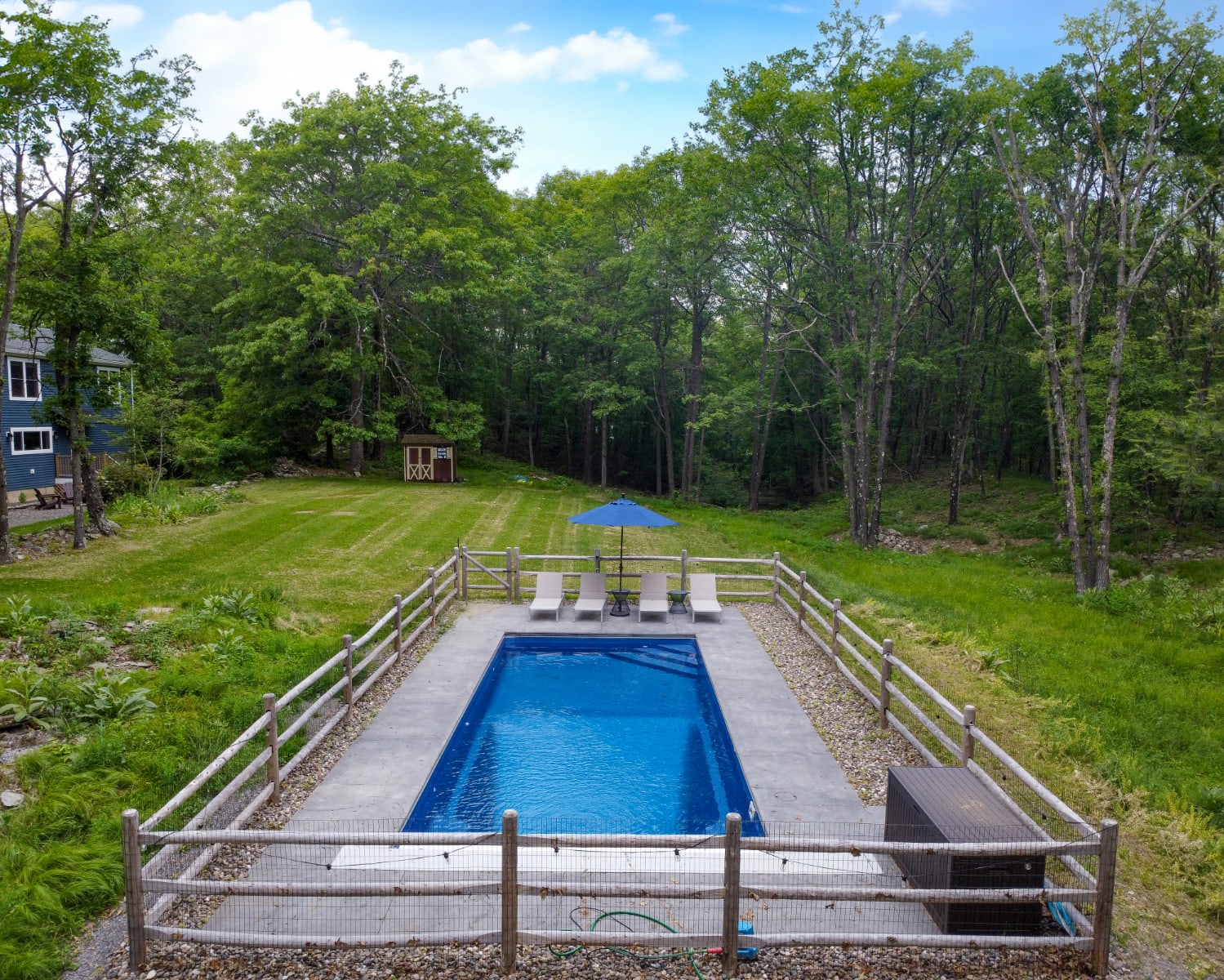An aerial view of a rectangular inground swimming pool, enclosed by a rustic wooden fence, with lounge chairs and an umbrella on the concrete surround. The pool is situated in a large grassy yard with a shed and dense forest in the background, and the house visible to the left.