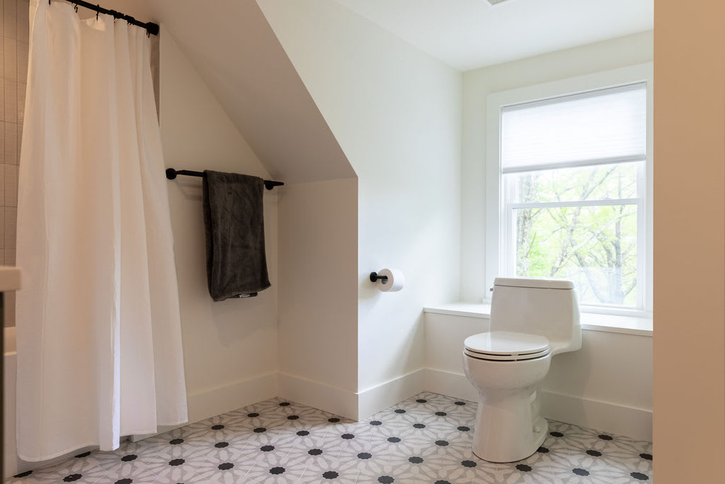 A bright bathroom corner showing a white toilet next to a large window, with a white shower curtain and a dark grey towel hanging on a black bar. The floor features a distinctive black and white patterned tile.