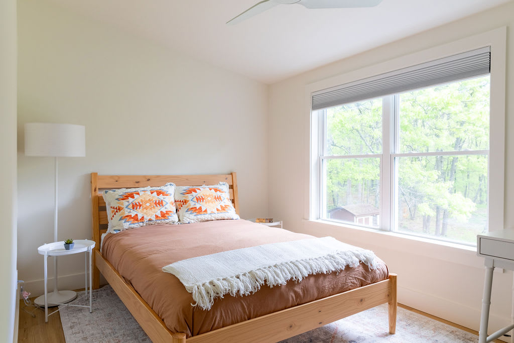 A cozy bedroom with a light wood bed frame, brown bedding, and decorative pillows with orange and blue patterns. A white floor lamp and a small round side table are next to the bed, with a large window offering views of trees.