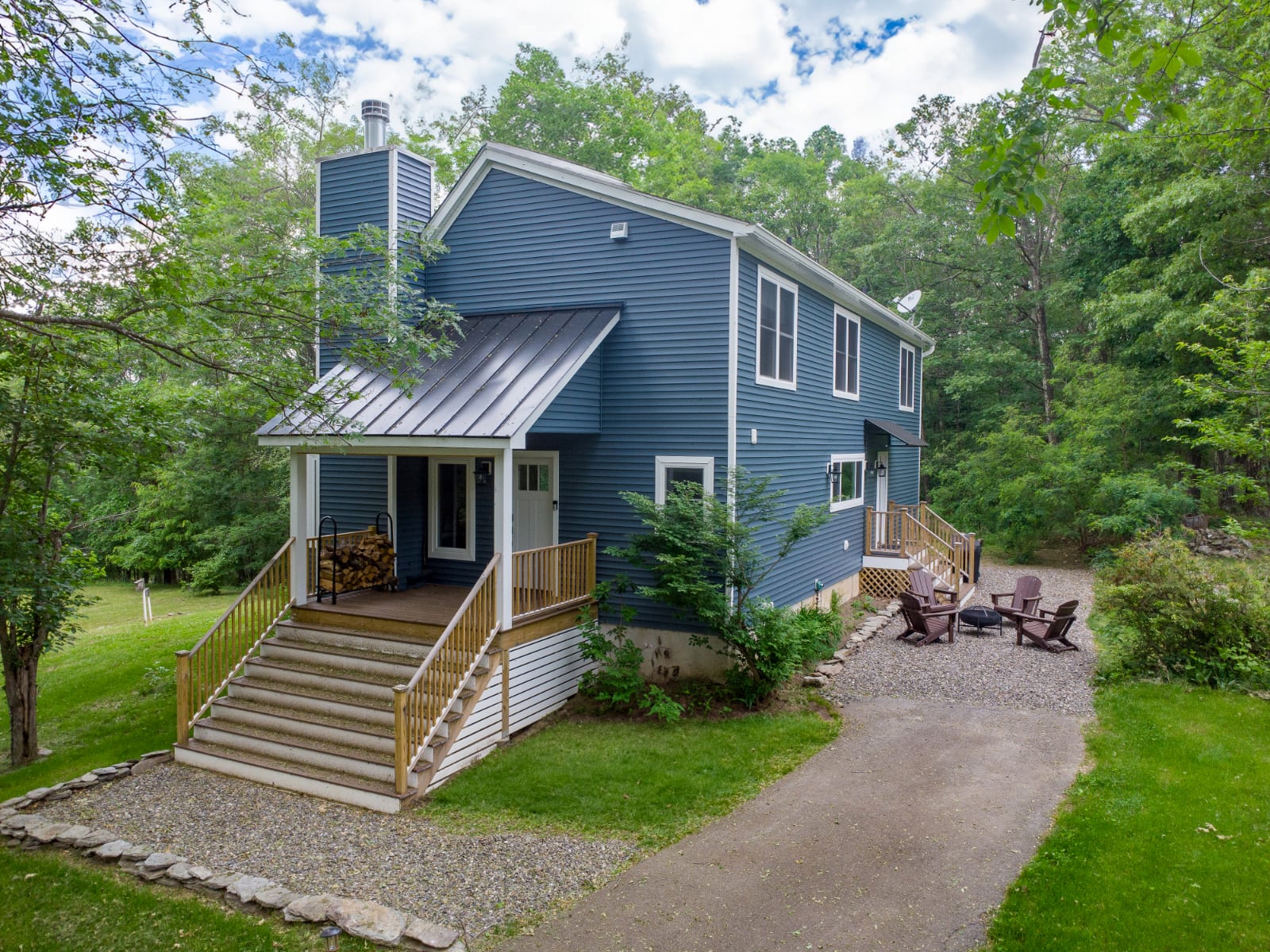 The front exterior of a two-story blue house with white trim and a dark metal roof, surrounded by lush green trees. A gravel driveway leads to the entrance, which features a wooden staircase and a small porch with stacked firewood.