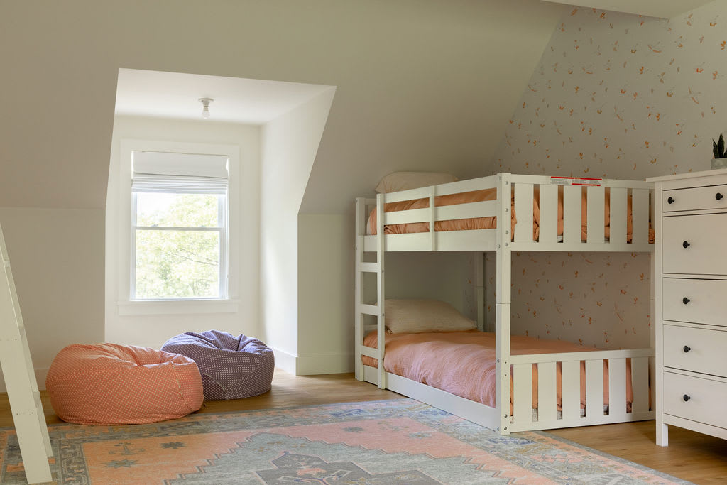 Angled view of a children's bedroom, showing one white bunk bed, a window with natural light, and two colorful beanbag chairs on a patterned rug. The room features sloped ceilings and patterned wallpaper.