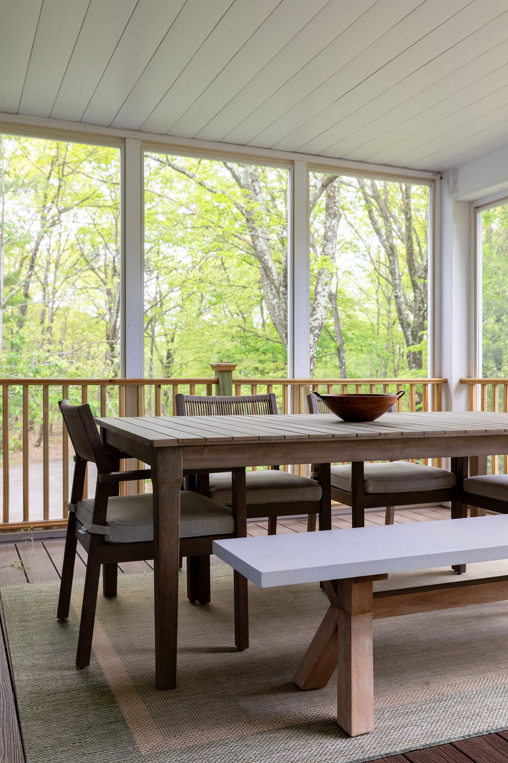 A light-filled screened porch features a wooden dining table with chairs and a bench, set on a woven rug, offering views of lush green trees through large windows.
