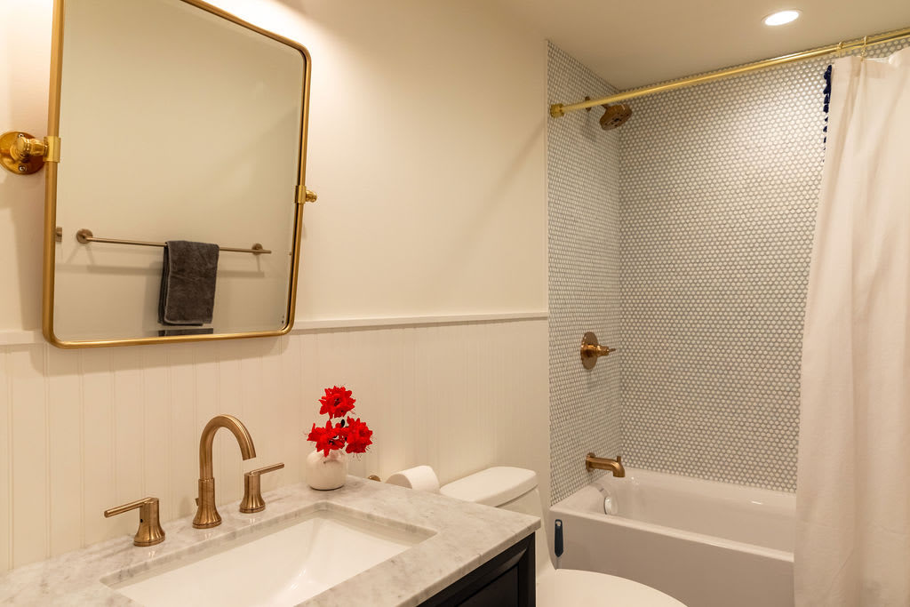 A bright bathroom featuring a dark vanity with a white marble countertop and gold fixtures, a rectangular gold-framed mirror, and a shower/tub combo with small white hexagonal tiles. White wainscoting lines the lower walls.
