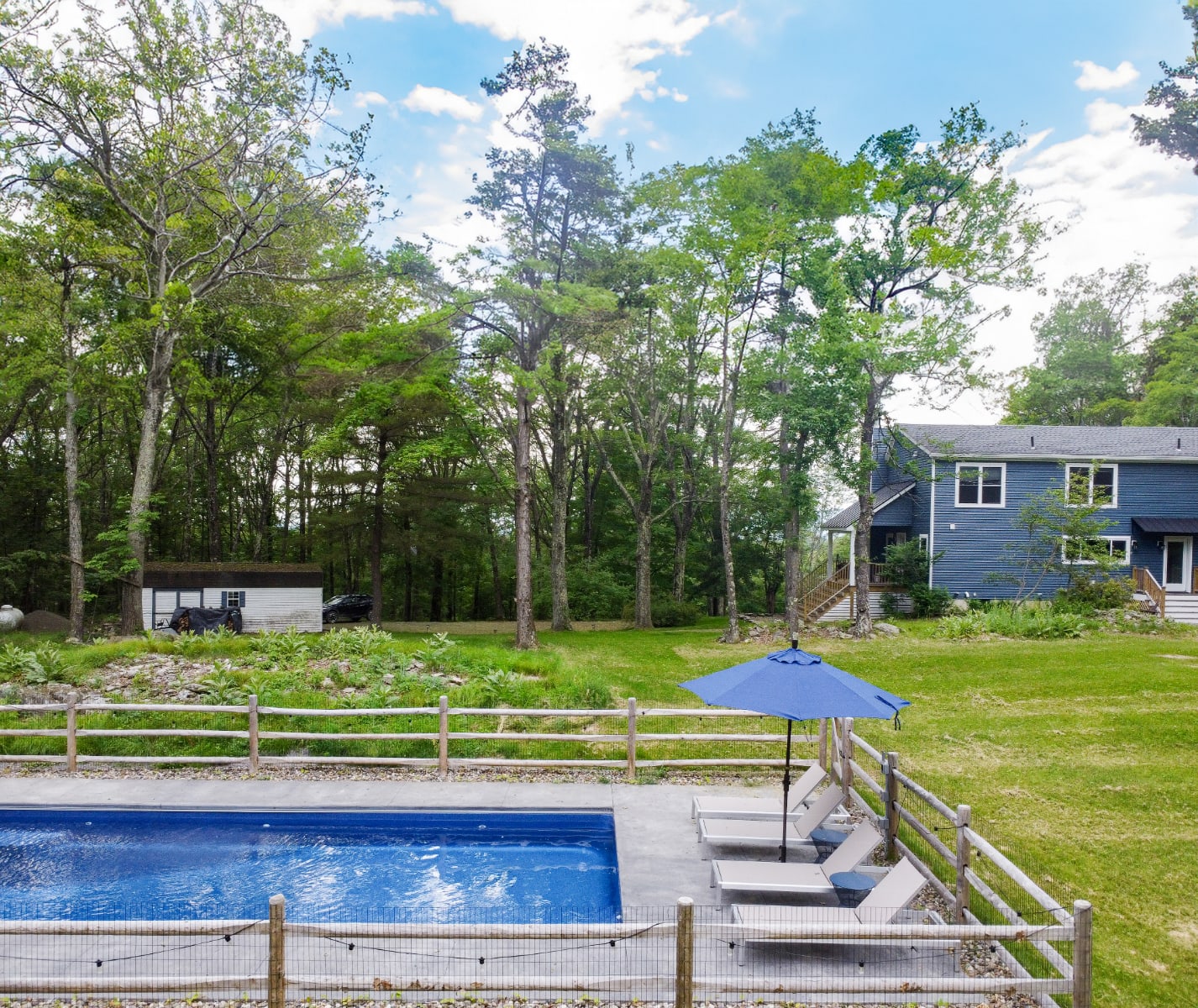 The rectangular outdoor swimming pool is surrounded by a concrete deck and a rustic wooden fence, with lounge chairs and a blue umbrella, and the blue house visible in the background amidst tall trees.