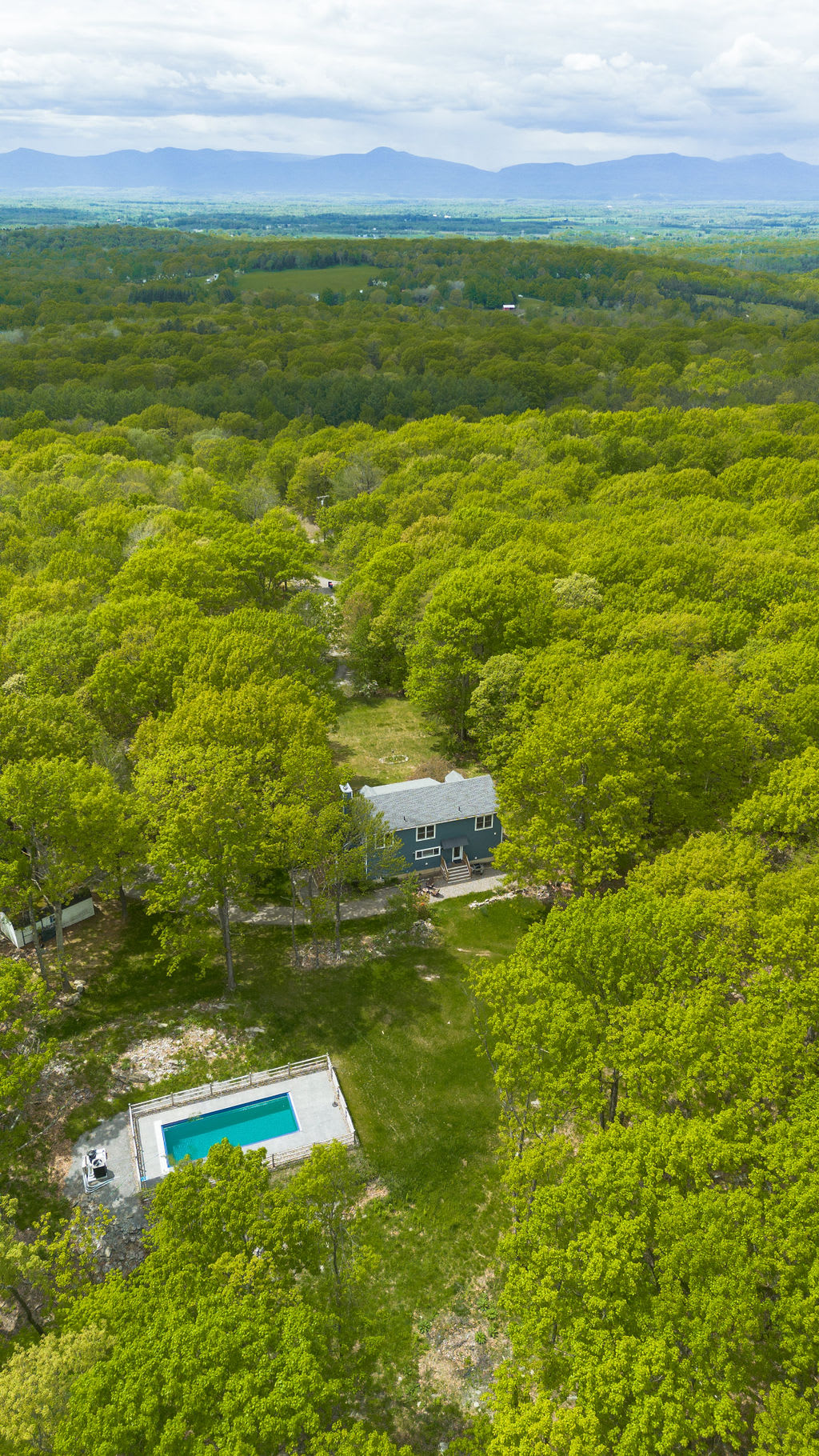 An aerial perspective shows the blue house and its rectangular outdoor pool nestled within a clearing of lush green forest, with distant mountains visible on the horizon under a bright sky.