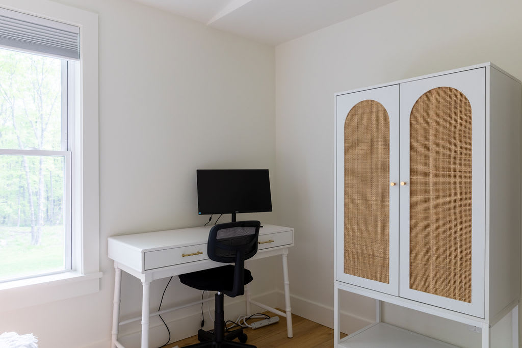 A dedicated workspace featuring a white desk with a black office chair and a computer monitor. A white and rattan cabinet stands nearby, and a large window provides natural light and a view of trees.