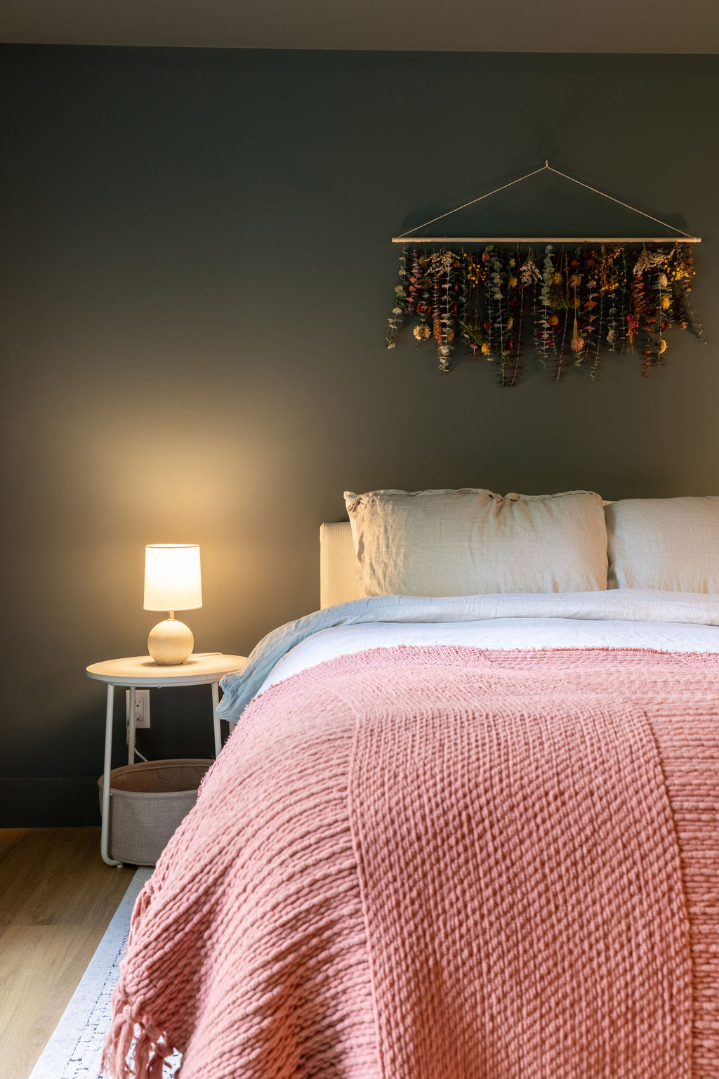 Cozy bedroom featuring a bed with a pink knitted blanket and neutral bedding, set against a dark green accent wall. A small white side table with a lamp provides soft lighting.