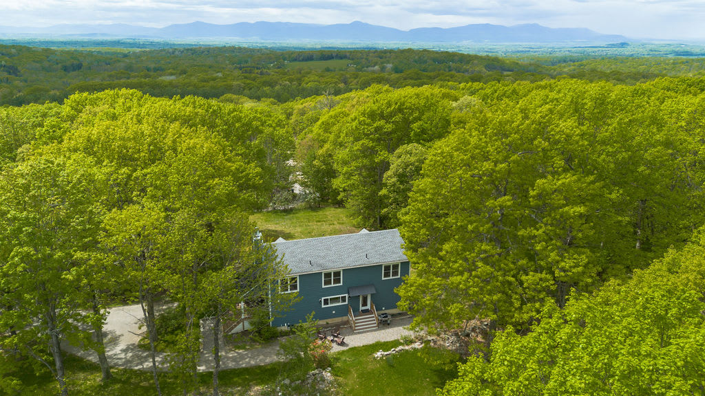 A high aerial view showcasing the blue house nestled among a vast expanse of lush green trees, with distant mountains visible on the horizon under a partly cloudy sky. The property appears secluded and surrounded by nature.