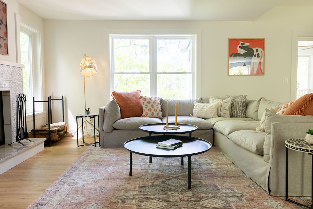 A slightly different angle of the bright living room, showcasing the large light gray sectional sofa, black coffee tables, and patterned rug. A tall woven floor lamp stands next to the sofa, and large windows offer views of the greenery outside.